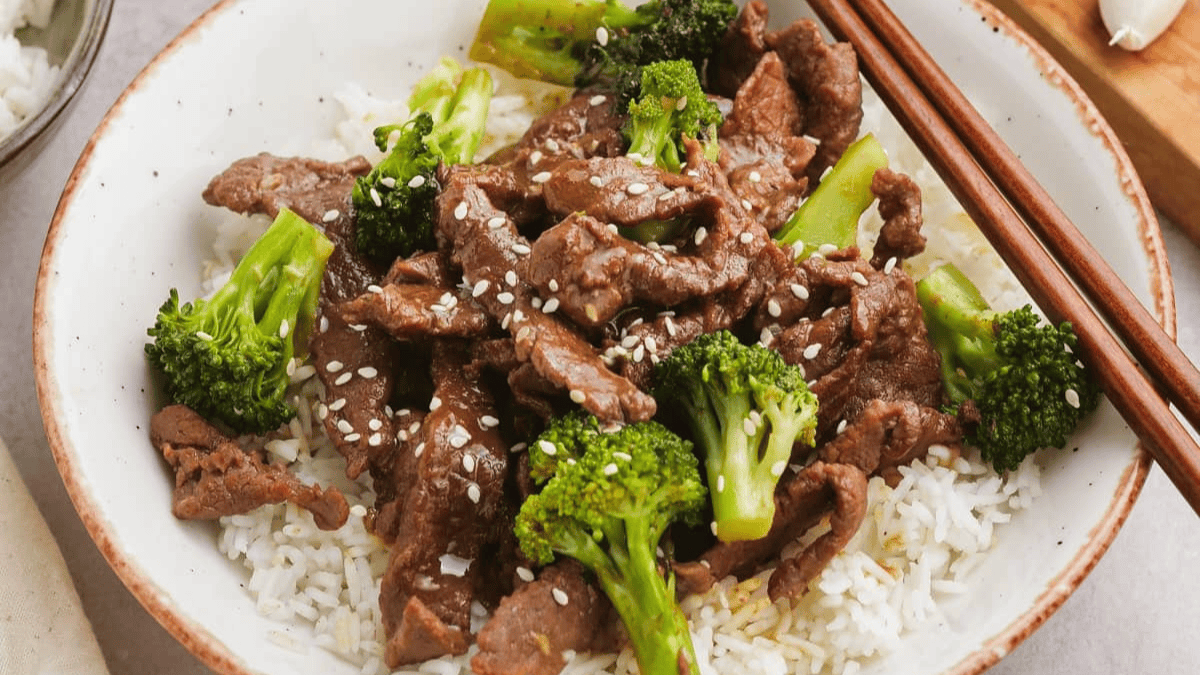 A bowl of white rice topped with beef stir-fry, broccoli florets, and sprinkled with sesame seeds, with a pair of wooden chopsticks resting on the side of the bowl.