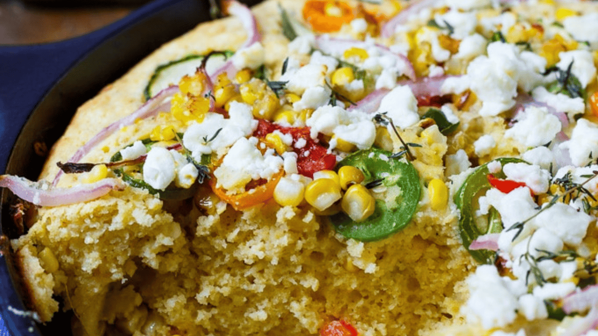 A close-up of cornbread topped with crumbled feta cheese, sliced jalapeños, red onions, corn kernels, and herbs, with a large piece removed to show the moist, fluffy texture inside.