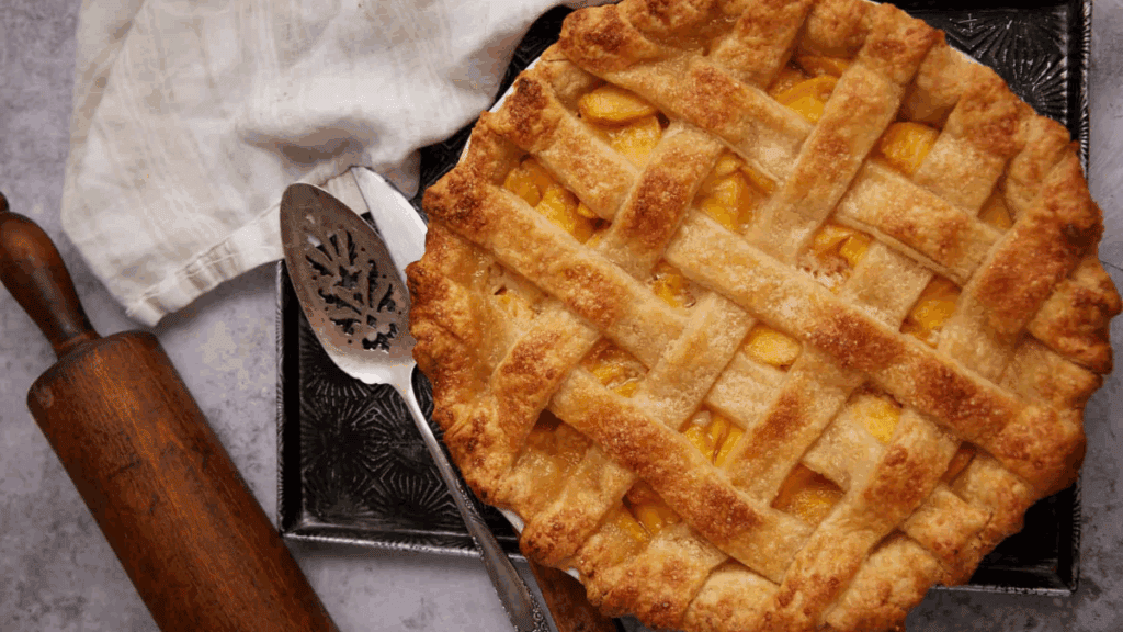 A golden-brown lattice-topped pie sits on a metal tray next to a rolling pin, a pie server, and a white cloth on a light surface.