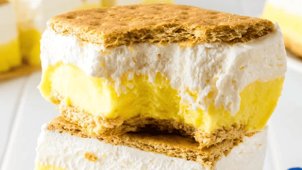 A close-up of two stacked yellow and white ice cream sandwiches with graham cracker cookies, one with a bite taken out, on a white surface.