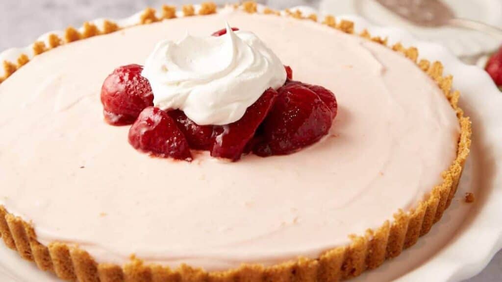 A creamy strawberry tart with a graham cracker crust, topped with glazed strawberries and a dollop of whipped cream, on a white plate.