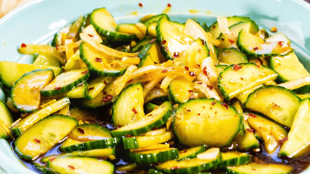 A close-up of a cucumber salad with thinly sliced cucumbers, onions, and red pepper flakes in a light, tangy dressing, served in a blue bowl.
