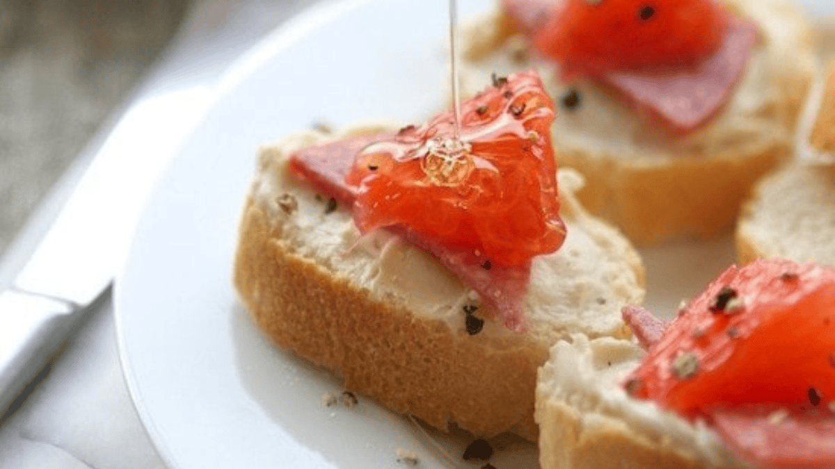 A close-up of sliced baguette topped with cream cheese, smoked salmon, black pepper, and a drizzle of honey being poured on top, served on a white plate.