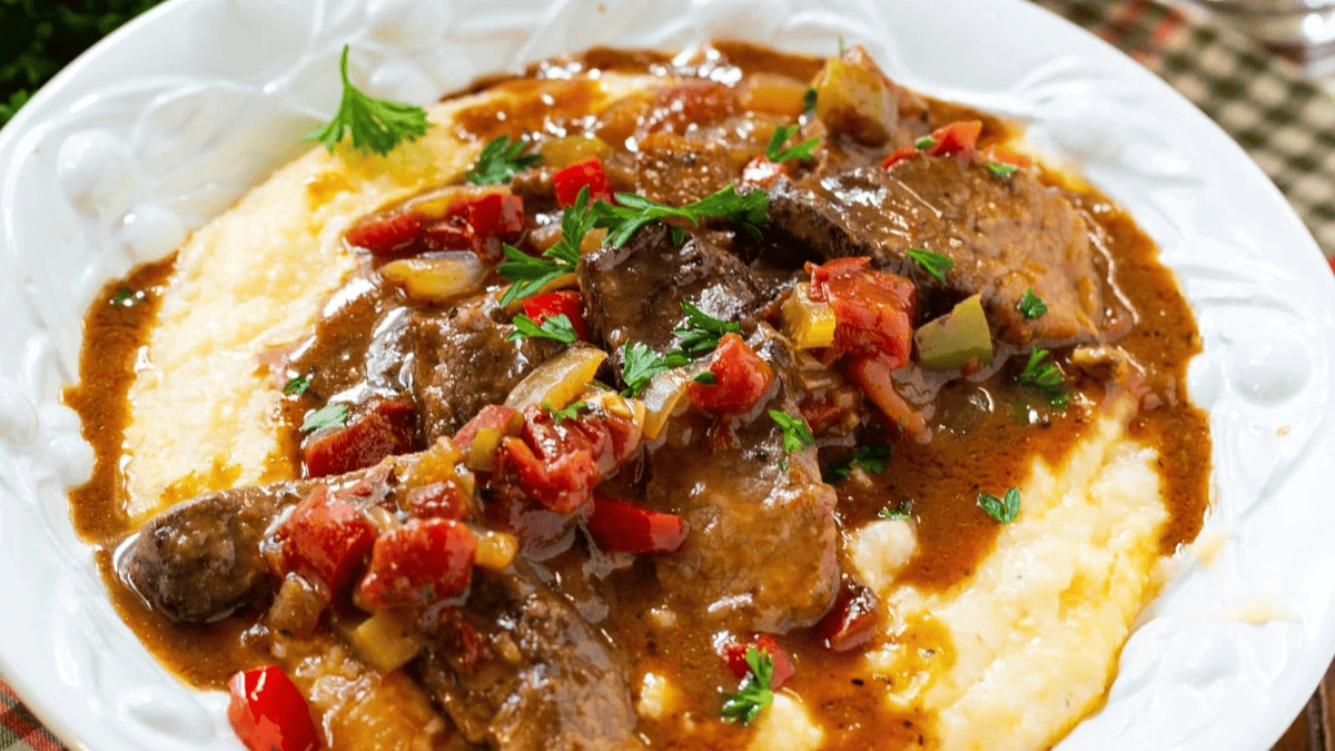 A plate of creamy yellow grits topped with slices of smothered beef, colorful diced tomatoes, peppers, celery, gravy, and garnished with chopped parsley.