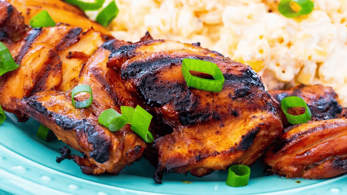Grilled pieces of chicken topped with sliced green onions on a blue plate, with a side of creamy macaroni and cheese in the background.