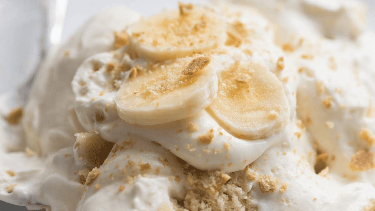 Close-up of banana pudding topped with whipped cream, banana slices, and crumbled cookies. The dessert appears creamy and smooth, with visible pieces of banana and cookie on top.