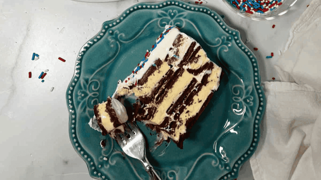 A slice of layered ice cream cake with chocolate and vanilla layers sits on an ornate teal plate, topped with white frosting and colorful sprinkles. A fork with a bite of cake rests beside the slice.