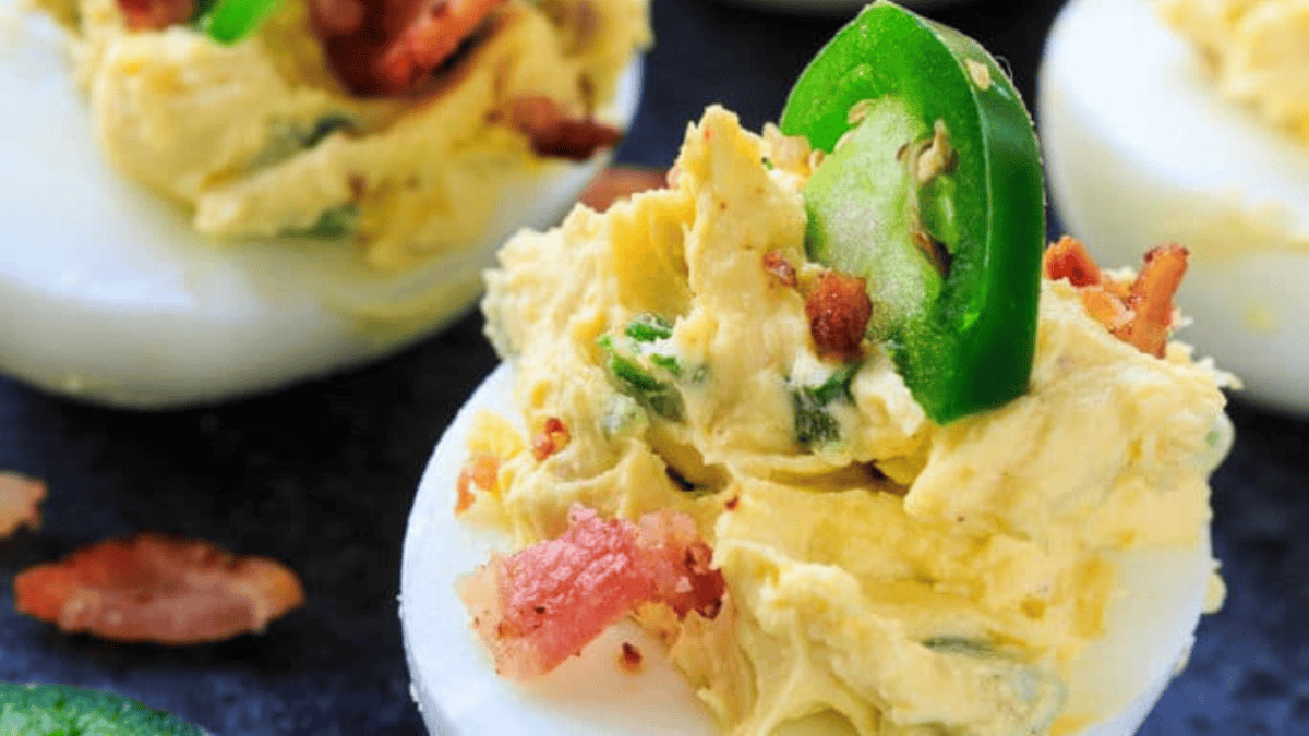 A close-up of deviled eggs topped with creamy yolk mixture, crispy bacon bits, and a slice of fresh jalapeño, set against a dark background.