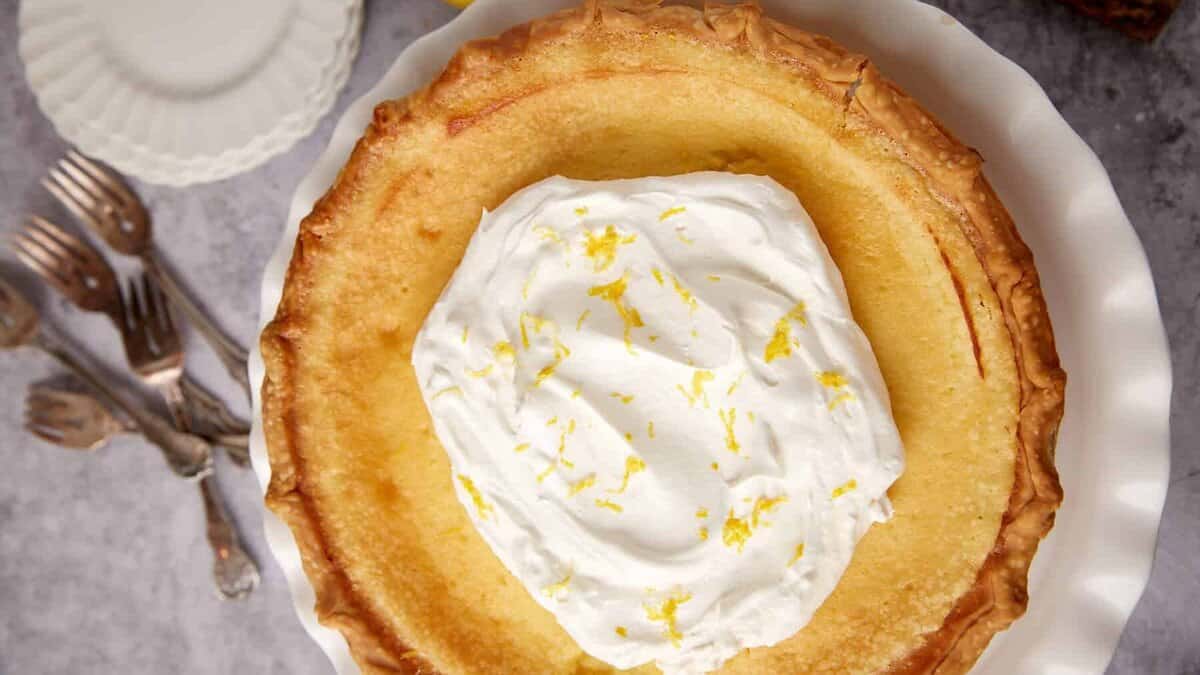 A lemon pie with a golden crust topped with whipped cream and lemon zest sits on a white plate, with forks and stacked plates in the background.