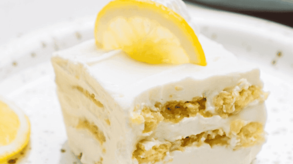 A square slice of layered lemon cake with creamy white frosting and a lemon wedge on top, served on a white plate.
