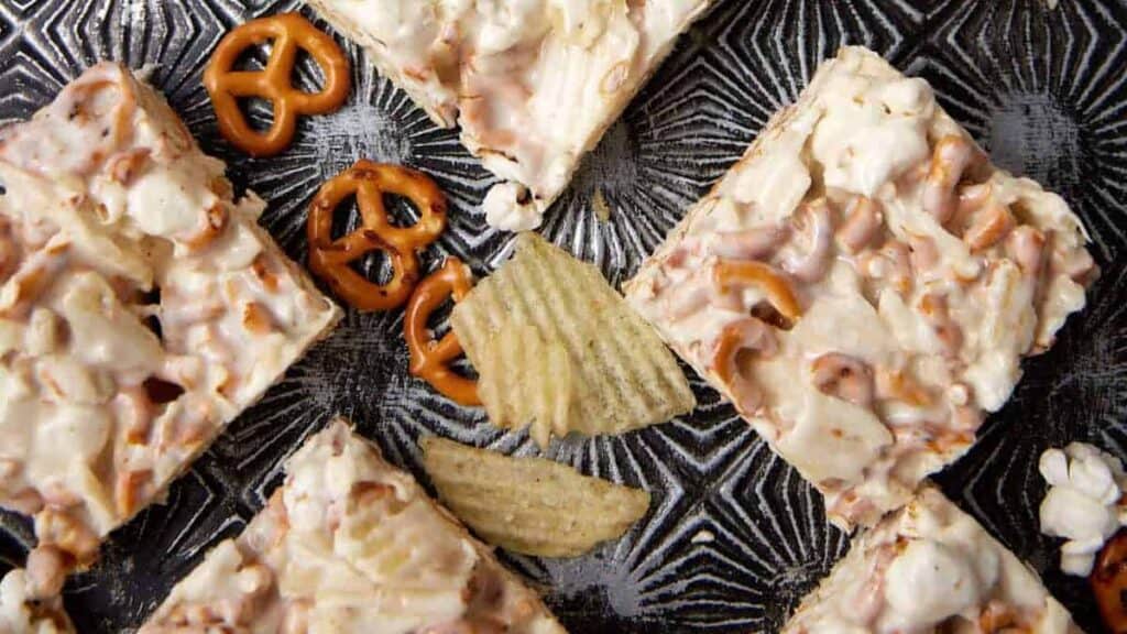 Four popcorn snack bars with pieces of pretzels and potato chips on a dark, patterned surface. Extra pretzels and wavy potato chips are scattered around the bars.