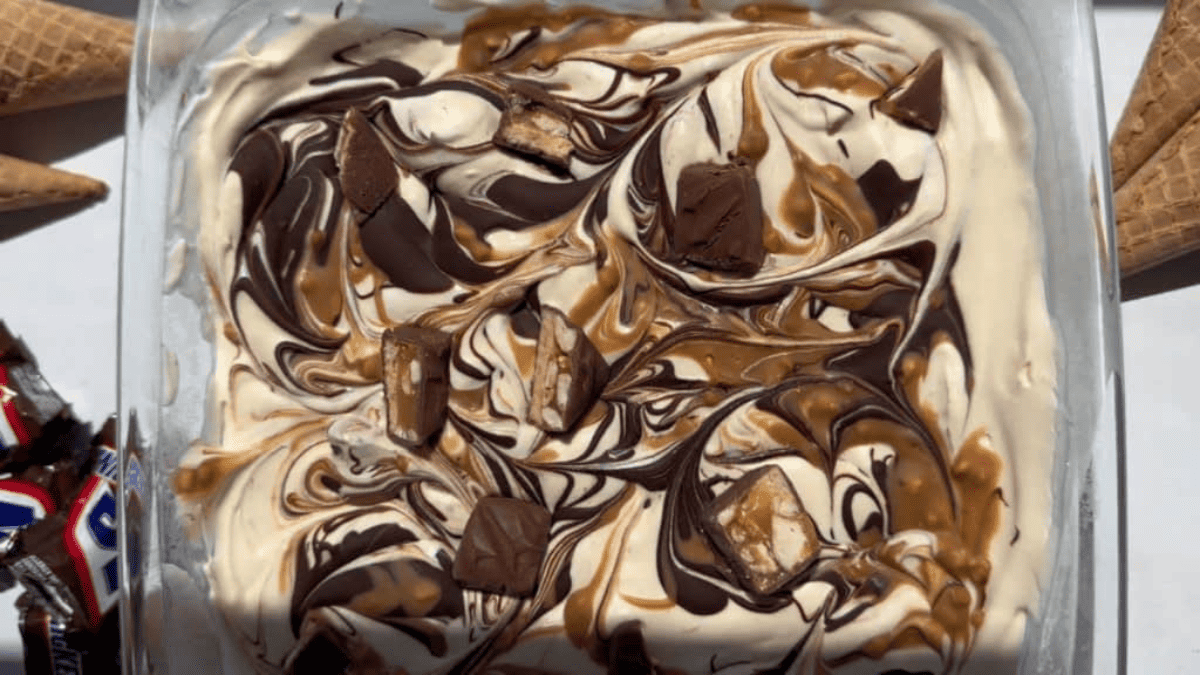 A rectangular dish of swirled chocolate and caramel ice cream topped with chocolate chunks. Waffle cones and candy bars are placed around the dish on a white surface.