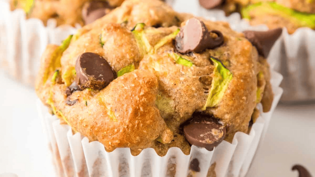 Close-up of a zucchini muffin with visible shreds of zucchini and several chocolate chips, in a white paper muffin liner.