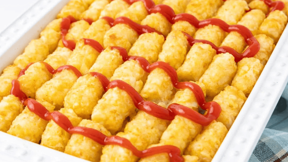 A white baking dish filled with rows of golden tater tots, topped with zigzag lines of ketchup.