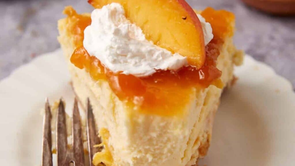 A slice of cheesecake topped with peach slices, peach glaze, and a dollop of whipped cream, served on a white plate with a fork beside it.