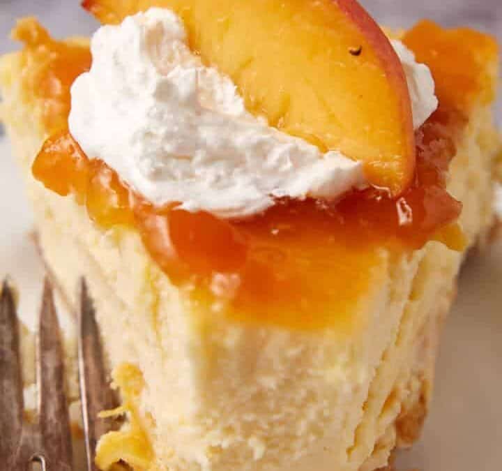 A slice of cheesecake topped with peach slices, peach glaze, and a dollop of whipped cream, served on a white plate with a fork beside it.