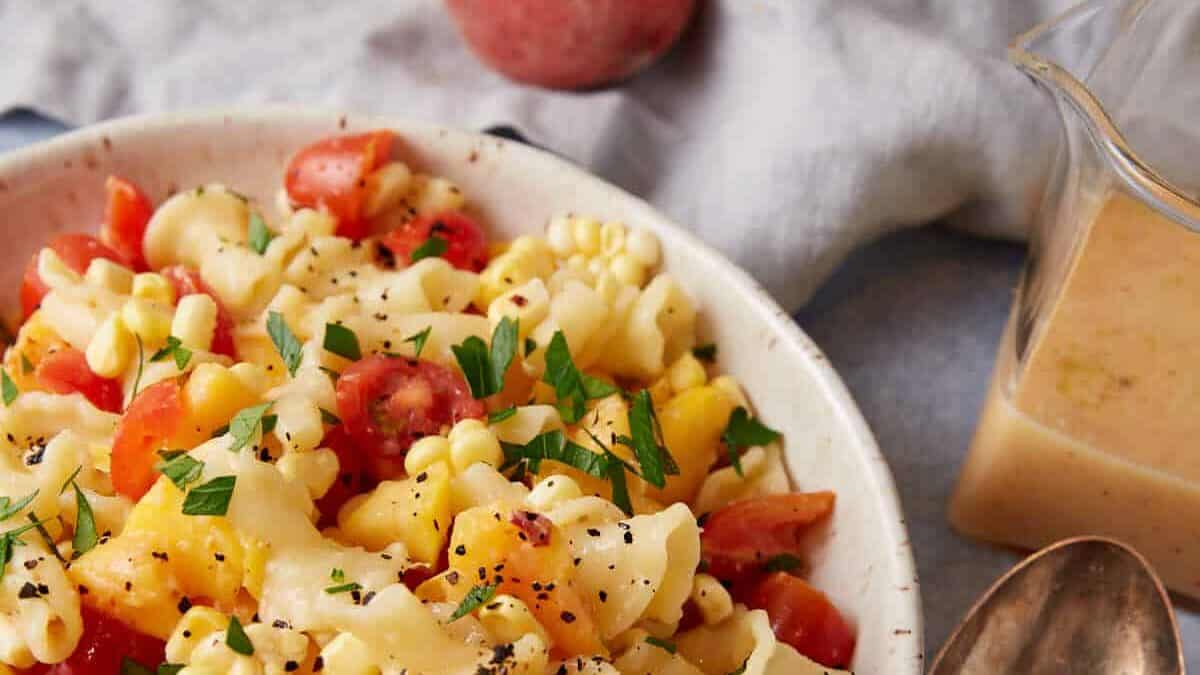 A bowl of peach pasta salad with dressing in a pticher on the side.