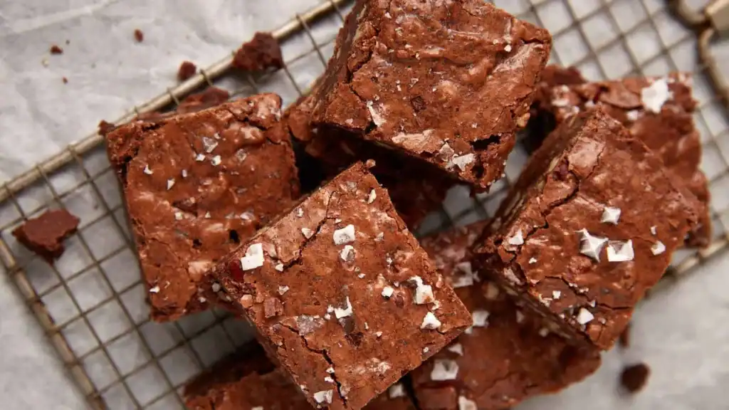 A close-up of several chocolate brownies topped with flaky sea salt, stacked on a wire cooling rack. The brownies have a crackly top and a rich, fudgy texture.