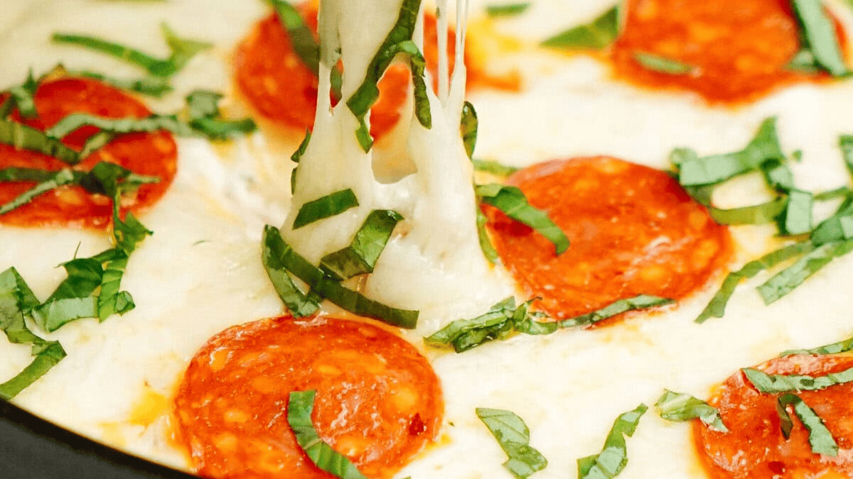 Close-up of melted cheese stretching as a slice is pulled from a pepperoni pizza dip, topped with fresh basil strips and round slices of pepperoni.