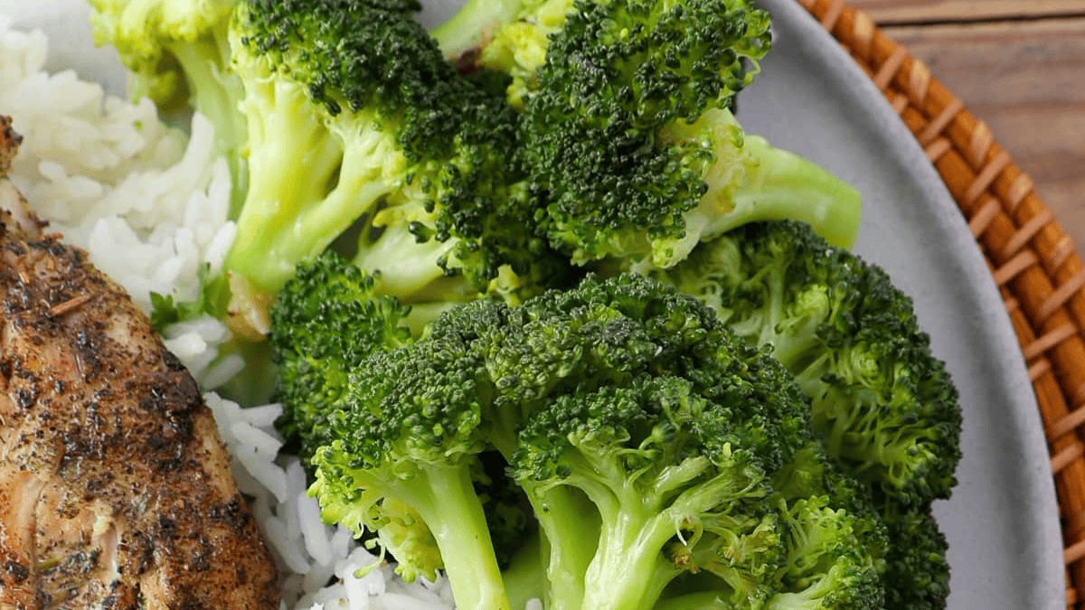 A plate with steamed broccoli florets and white rice, partially visible grilled chicken on the side, served on a gray dish with a wicker placemat underneath.