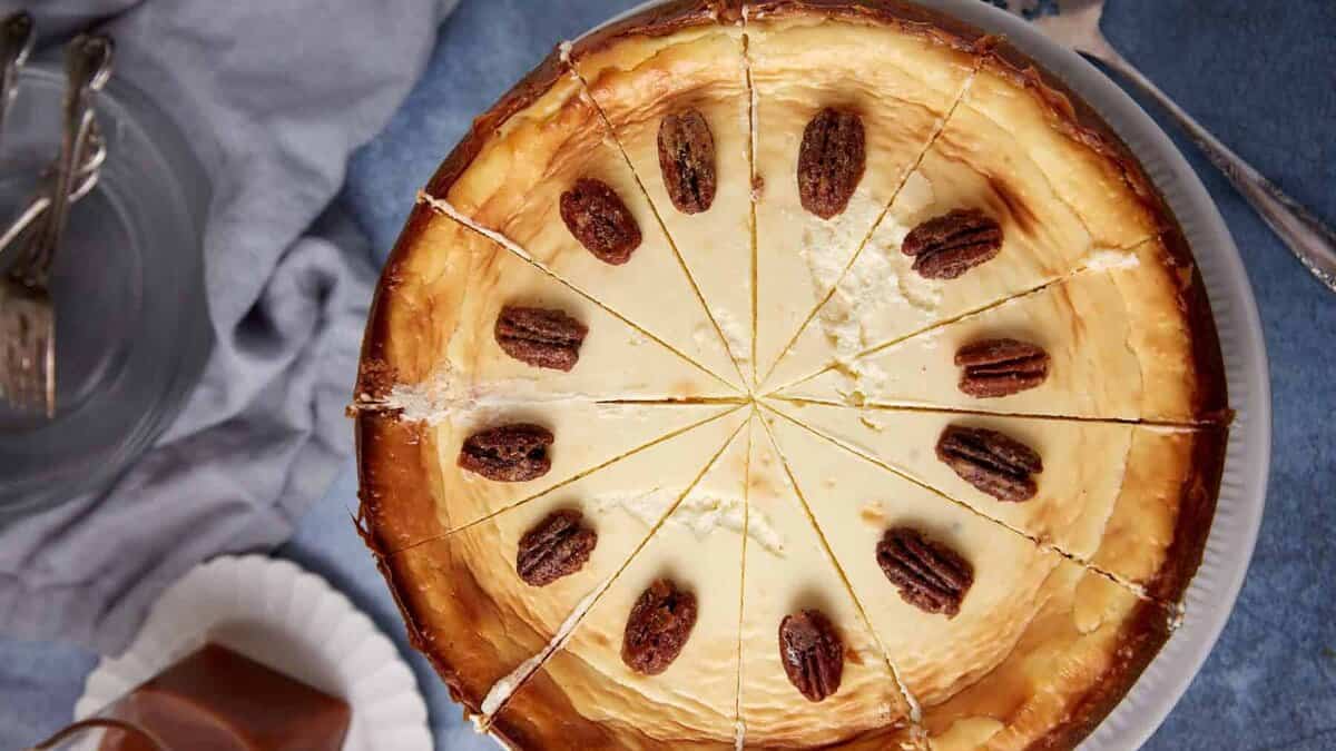 A top view of a baked cheesecake sliced into 12 pieces, each topped with a pecan half, on a white plate. A fork, plates, and a napkin are visible in the background.