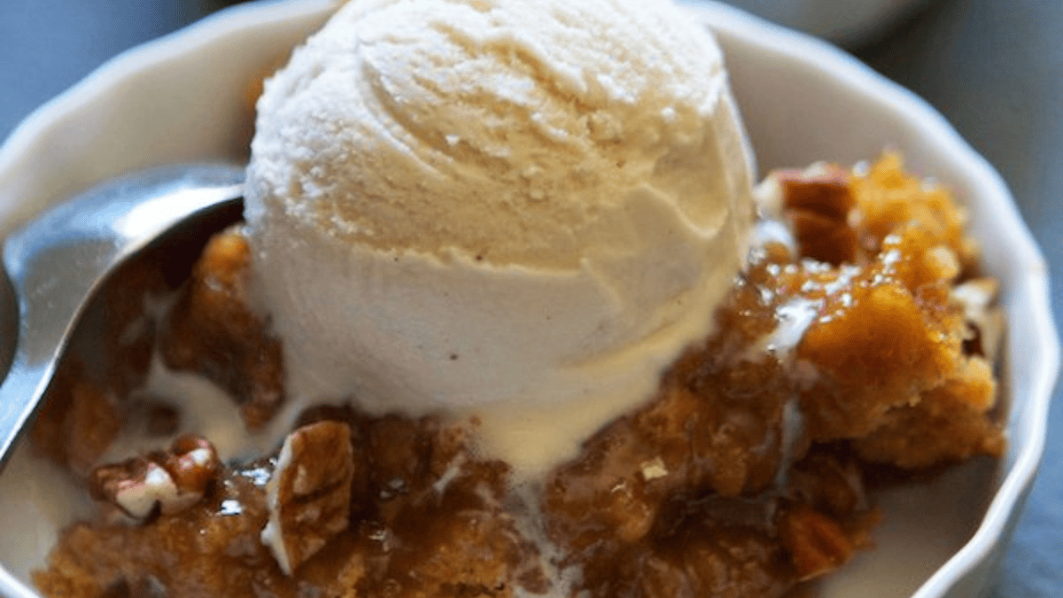 A bowl of warm dessert topped with a scoop of vanilla ice cream, pecan pieces, and a spoon resting on the side.