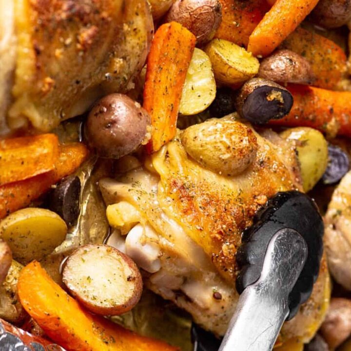 Tongs in a pan of baked chicken with carrots and potatoes.