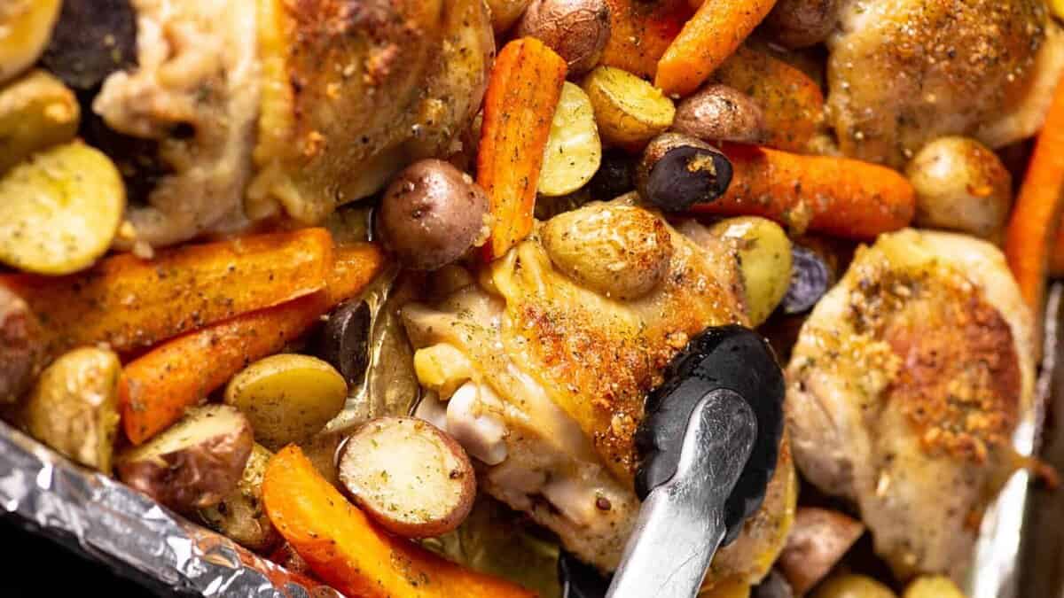Tongs in a pan of baked chicken with carrots and potatoes.
