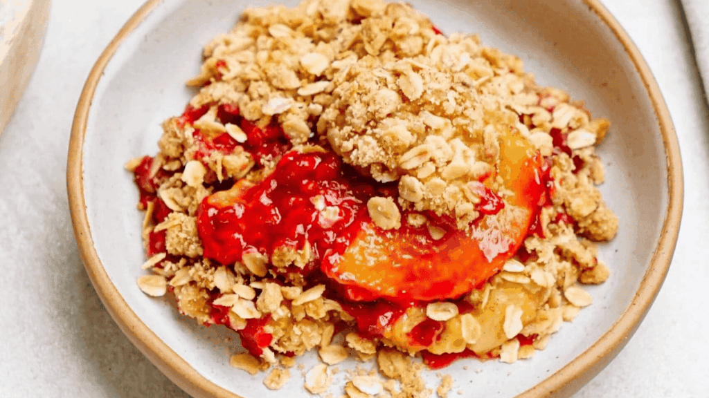 A close-up of a serving of fruit crumble dessert on a white plate, featuring golden oat topping and bright red and yellow fruit filling.