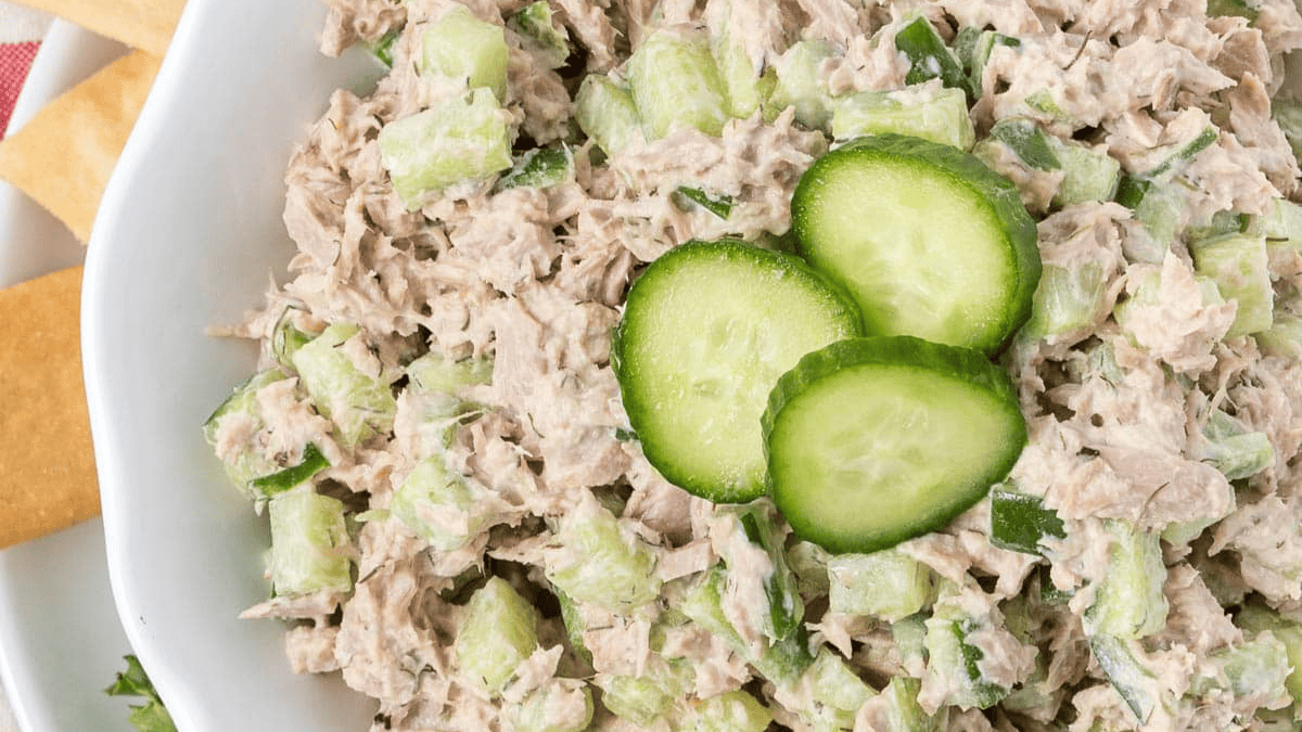 A bowl of tuna salad mixed with chopped celery and herbs, garnished with three slices of cucumber on top. Crackers are visible on the side of the bowl.