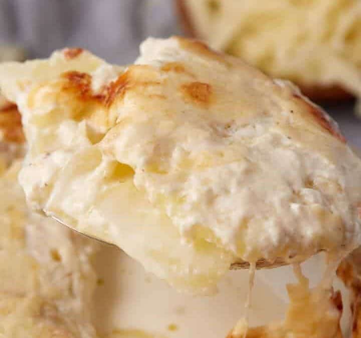 A close-up of a spoon lifting a cheesy, creamy serving of scalloped potatoes with cream from a dish, with melted cheese on top and a small pile of shredded cheese in the background.