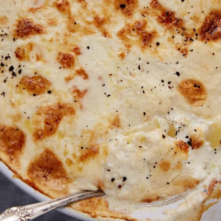 A close-up of homemade scalloped potatoes with a golden, bubbly cheese and cream topping, sprinkled with black pepper. A serving spoon rests in the dish, and a small bowl of grated cheese is visible in the background.