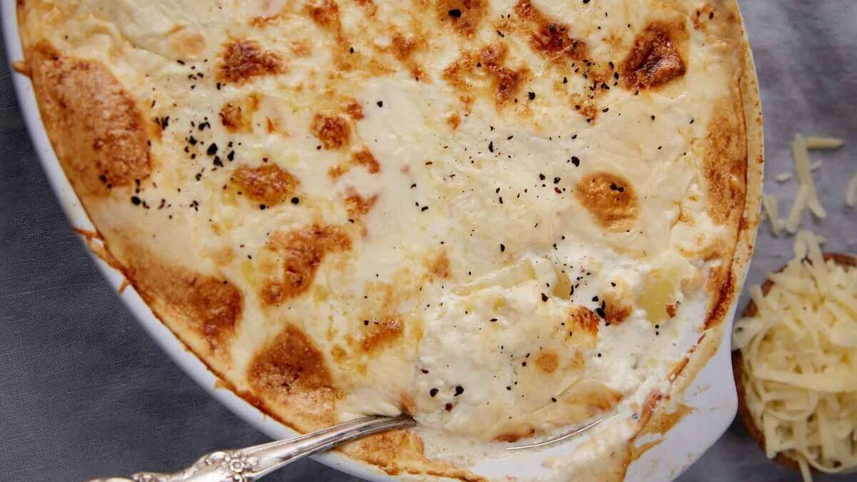 A close-up of homemade scalloped potatoes with a golden, bubbly cheese and cream topping, sprinkled with black pepper. A serving spoon rests in the dish, and a small bowl of grated cheese is visible in the background.