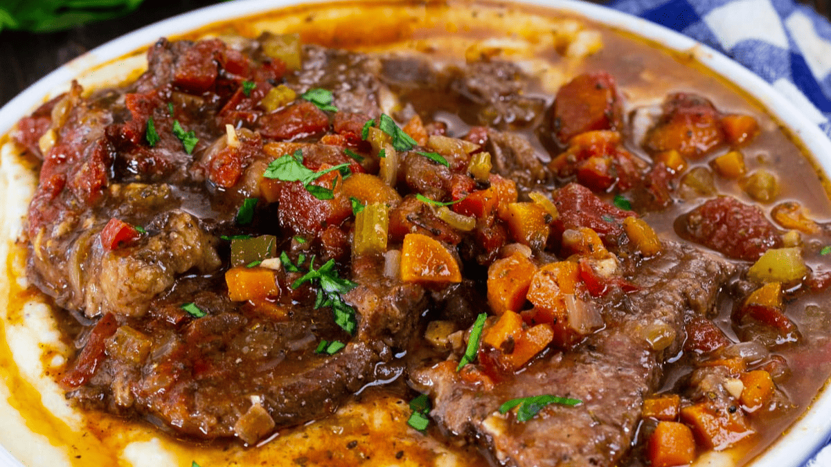 A plate of braised beef topped with a chunky tomato, carrot, and herb sauce, served over creamy mashed potatoes. The dish is garnished with chopped fresh parsley.