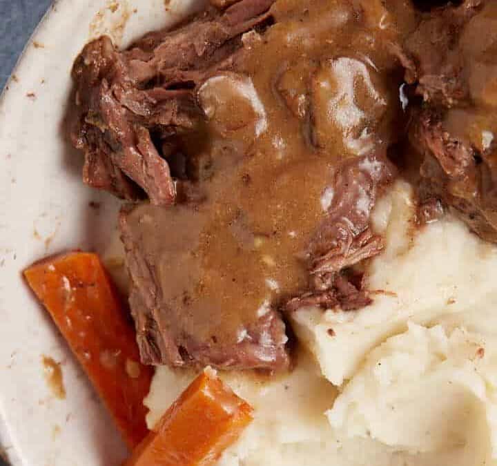 A white bowl filled with mashed potatoes, tender slow cooker pot roast with gravy, a carrot piece, all covered in brown gravy. A fork and knife are placed on a napkin next to the bowl, and a container of additional gravy is seen in the background.