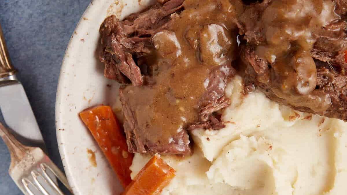 A white bowl filled with mashed potatoes, tender slow cooker pot roast with gravy, a carrot piece, all covered in brown gravy. A fork and knife are placed on a napkin next to the bowl, and a container of additional gravy is seen in the background.
