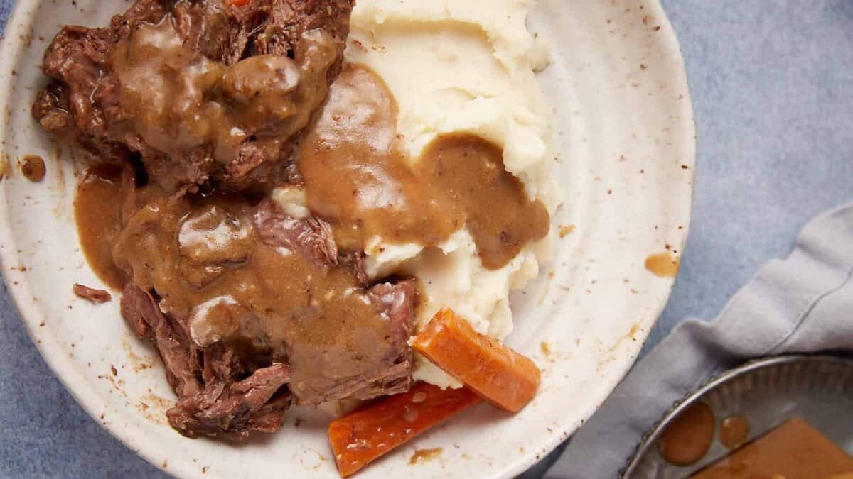A plate of mashed potatoes topped with brown gravy, accompanied by slow-cooked beef and roasted carrot sticks. There is a fork and knife on the right side of the plate, and a metal jug of gravy in the background on a cloth napkin. This hearty meal features savory slow cooker pot roast with gravy.