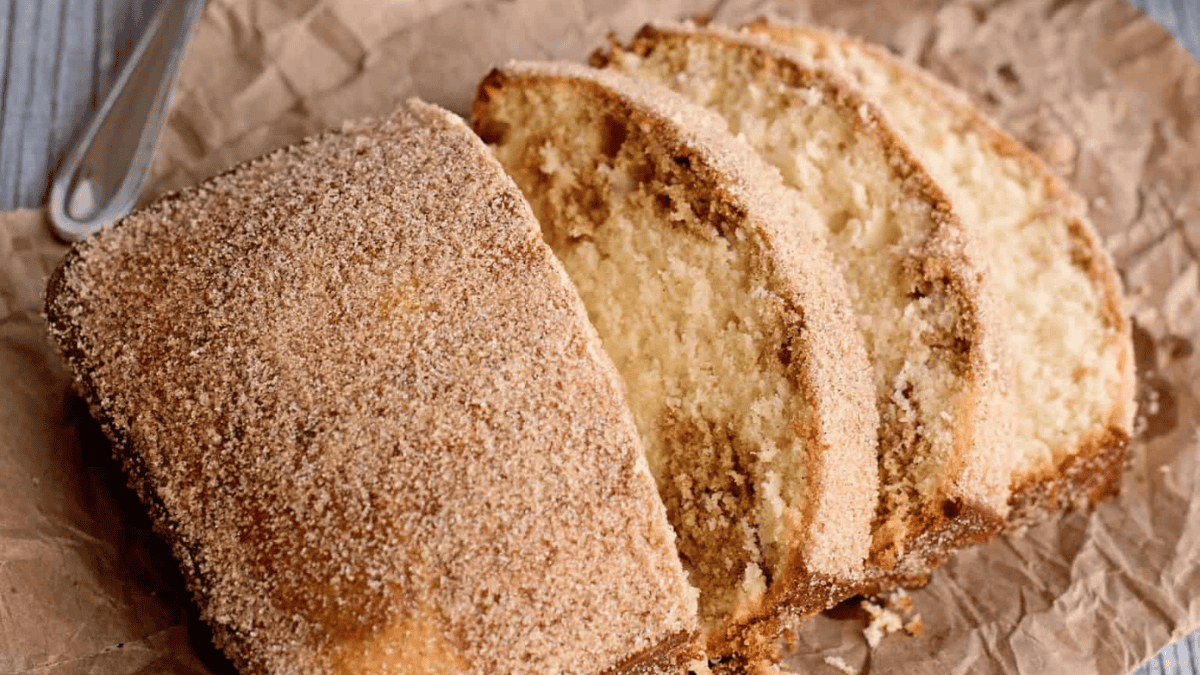 A loaf of cinnamon sugar bread sits on crumpled parchment paper, with three thick slices cut and fanned out to show the light, moist interior and a generous cinnamon sugar coating on top.