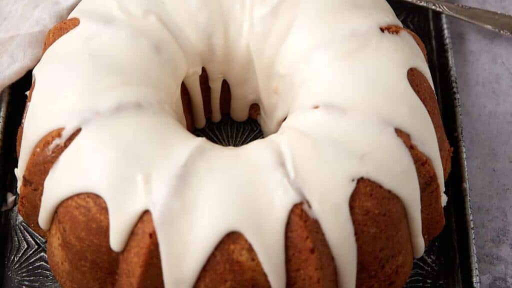 A Bundt cake with thick white icing drizzled over the top, sitting on a dark tray.