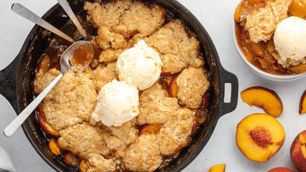 A skillet peach cobbler topped with two scoops of vanilla ice cream, served with two spoons. A bowl of cobbler with ice cream and sliced fresh peaches are placed nearby on a light surface.