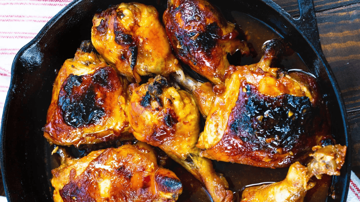 Chicken drumsticks and thighs with golden-brown, slightly charred skin, coated in a sticky glaze, arranged in a black cast iron skillet.