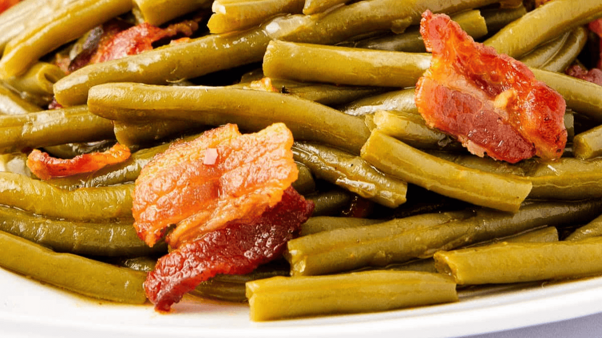 Close-up of cooked green beans mixed with crispy bacon pieces on a white plate.