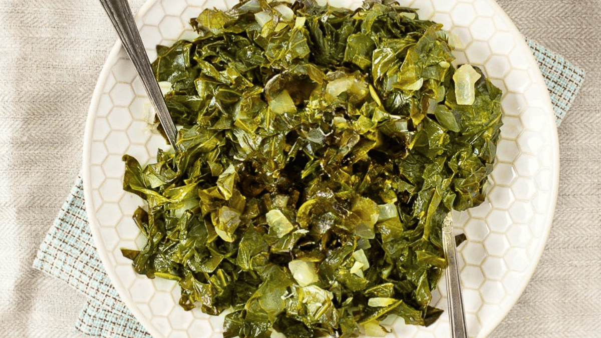 A white plate filled with cooked collard greens and chopped onions, with two serving spoons placed on either side of the dish. The plate rests on a textured cloth.