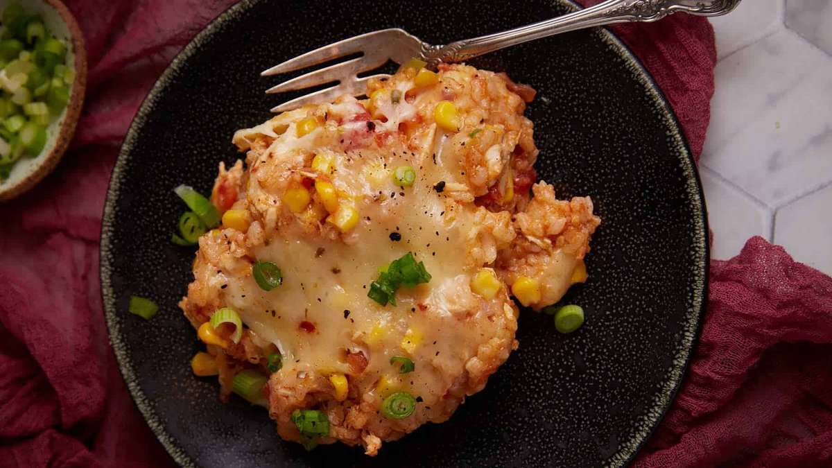 A black plate holds a serving of cheesy baked rice topped with melted cheese, corn, chopped green onions, and black pepper, with a silver fork resting on top. A maroon cloth and a small bowl are in the background.