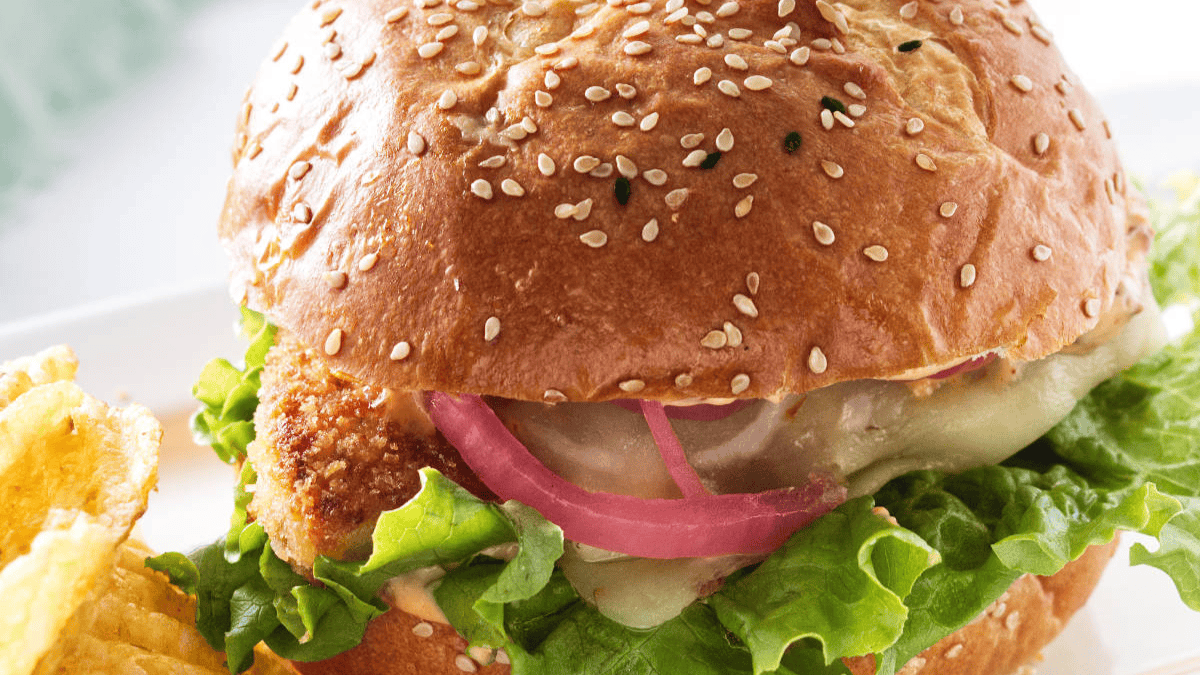 A close-up of a sesame seed bun sandwich filled with leafy green lettuce, melted cheese, pickled red onions, and a breaded patty, served next to a pile of potato chips.