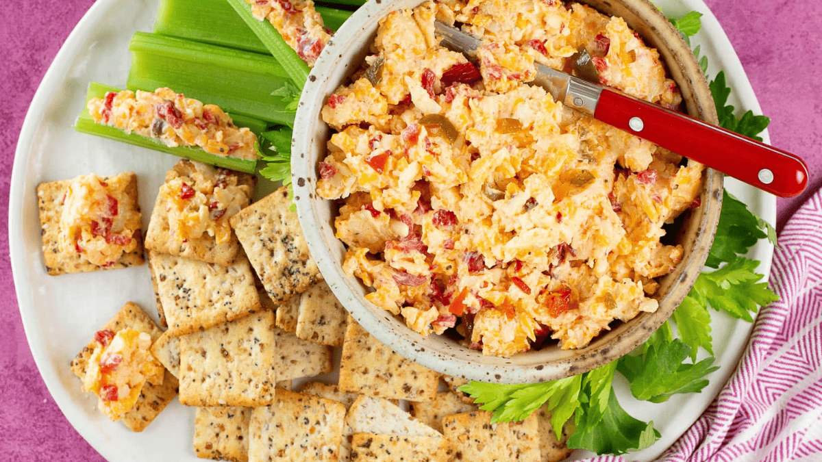 A bowl of pimento cheese dip with a red-handled spreader, surrounded by celery sticks and assorted crackers on a white plate, set on a pink surface.