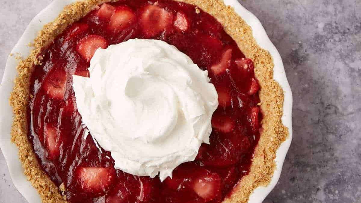 A strawberry pie with a graham cracker crust, topped with a large swirl of whipped cream, sits on a gray textured surface.