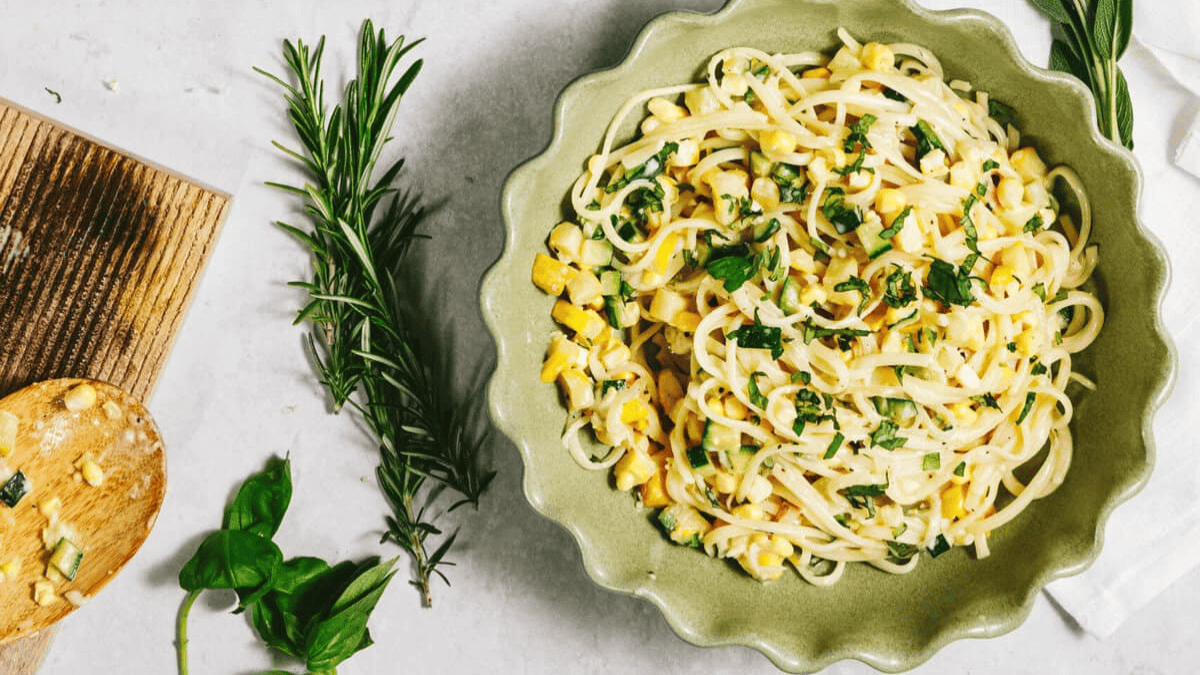 A scalloped green bowl filled with pasta mixed with corn and chopped herbs sits on a light surface beside fresh rosemary, basil, and a wooden spoon with some leftover corn.