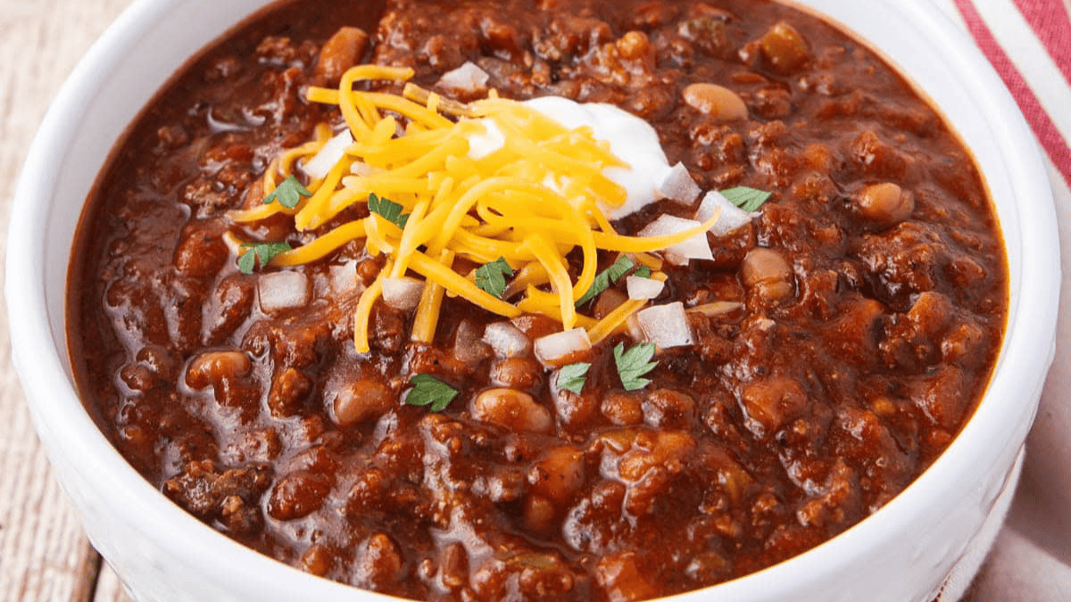 A bowl of chili topped with shredded cheddar cheese, a dollop of sour cream, diced onions, and fresh herbs. The chili looks thick with beans and meat, and is served in a white bowl on a wooden surface.