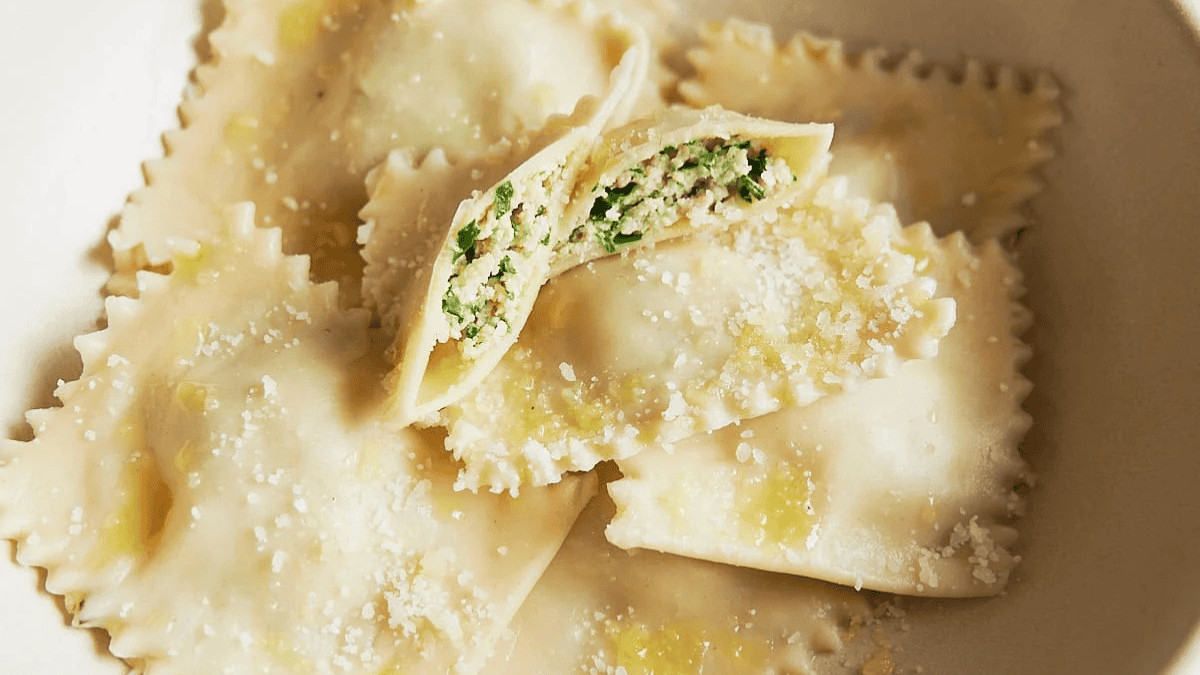 Close-up of ravioli topped with grated cheese and a drizzle of olive oil, with one ravioli cut open to show a green and white filling, all served in a white bowl.